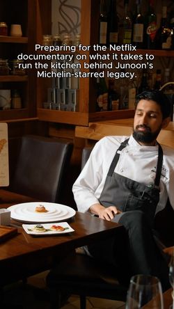 Chef in kitchen attire seated by a table with plated Indian cuisine dishes in an upscale restaurant setting.