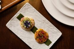 Two elegant Indian appetizers served on a textured white rectangular plate with a green leaf beneath each item.