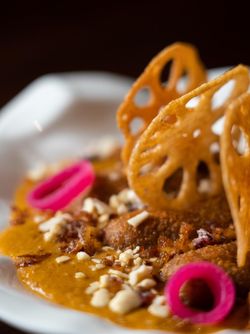 Close-up of an Indian curry dish garnished with nuts, pickled onions, and lotus root chips on a white plate.