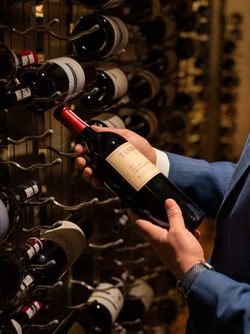 Person in blue suit selecting a red wine bottle from a dimly lit wine rack in an upscale Indian restaurant.