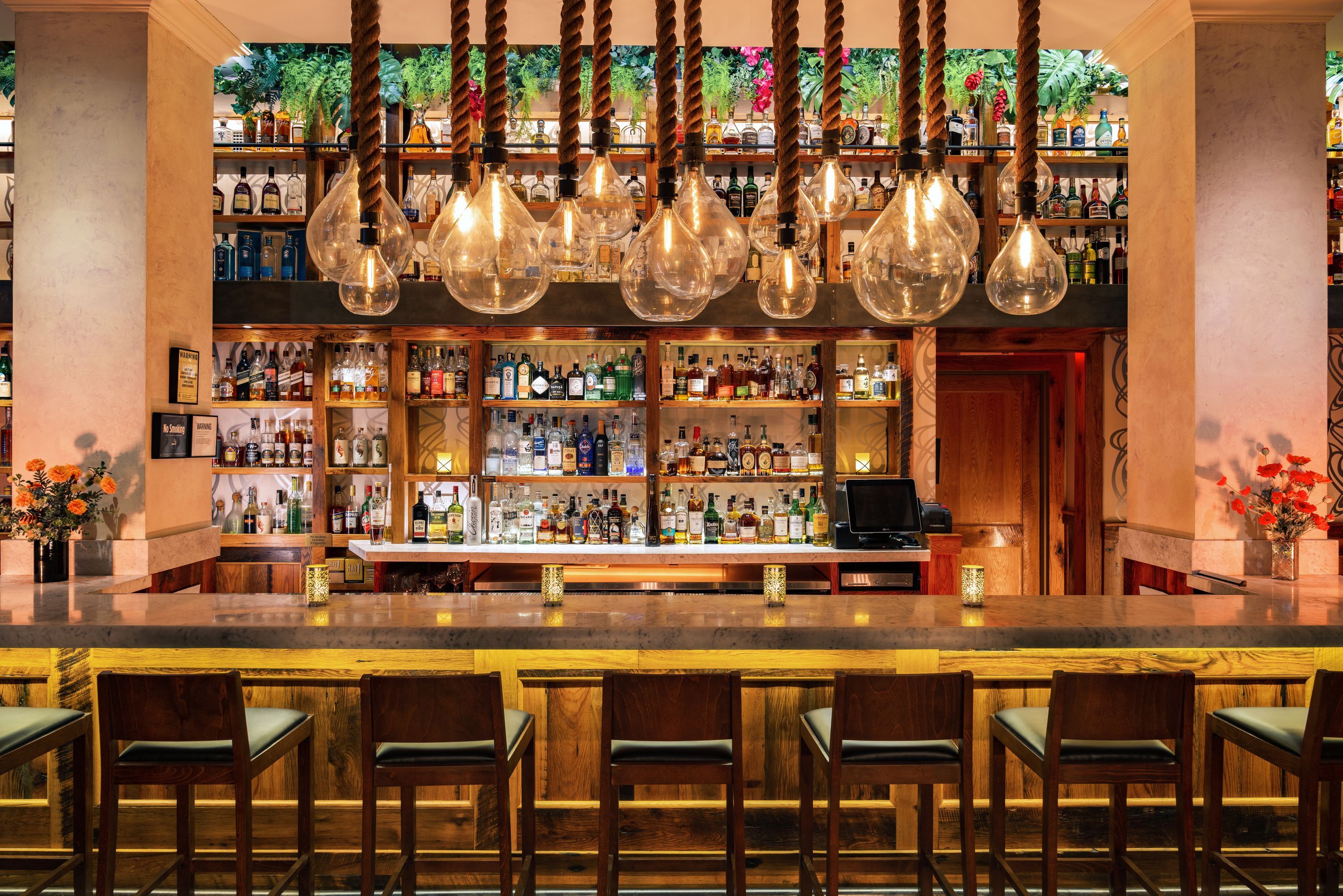 Bar area with hanging light bulbs, wooden bar counter, high chairs, and shelves stocked with various bottles.