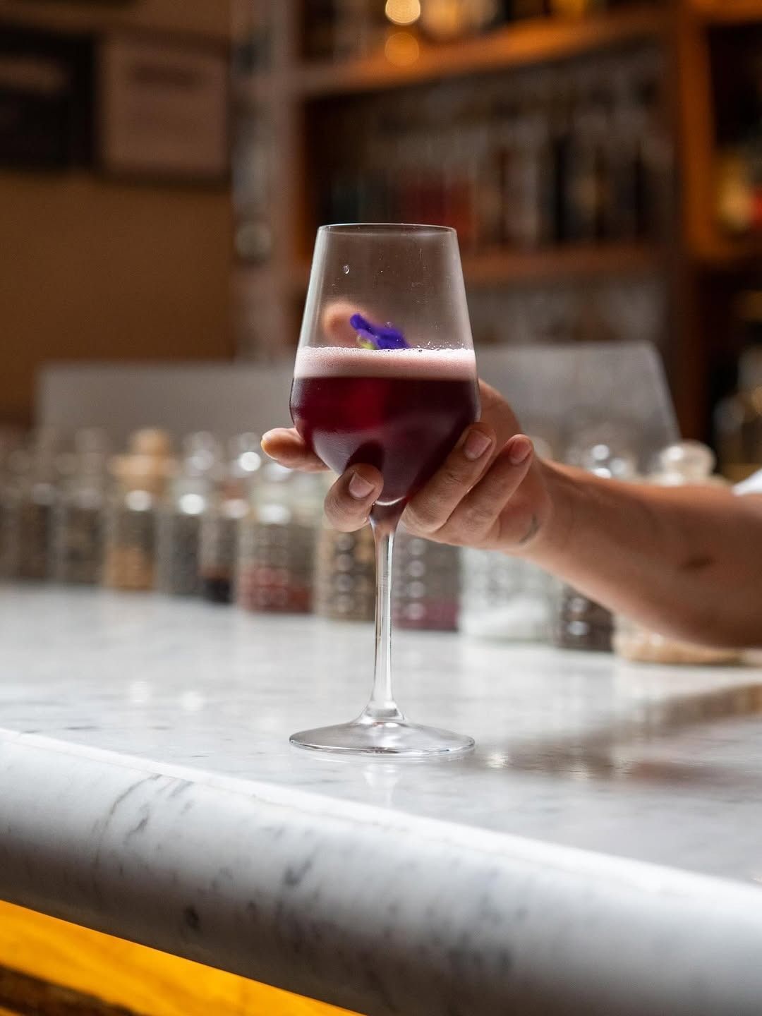 A hand holding a garnished red cocktail in a wine glass on a marble bar counter at an upscale Indian restaurant.
