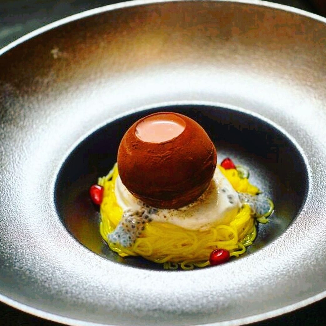 Fine dining Indian restaurant dessert featuring delicate yellow noodles, creamy sauce, and a chocolate sphere on top.
