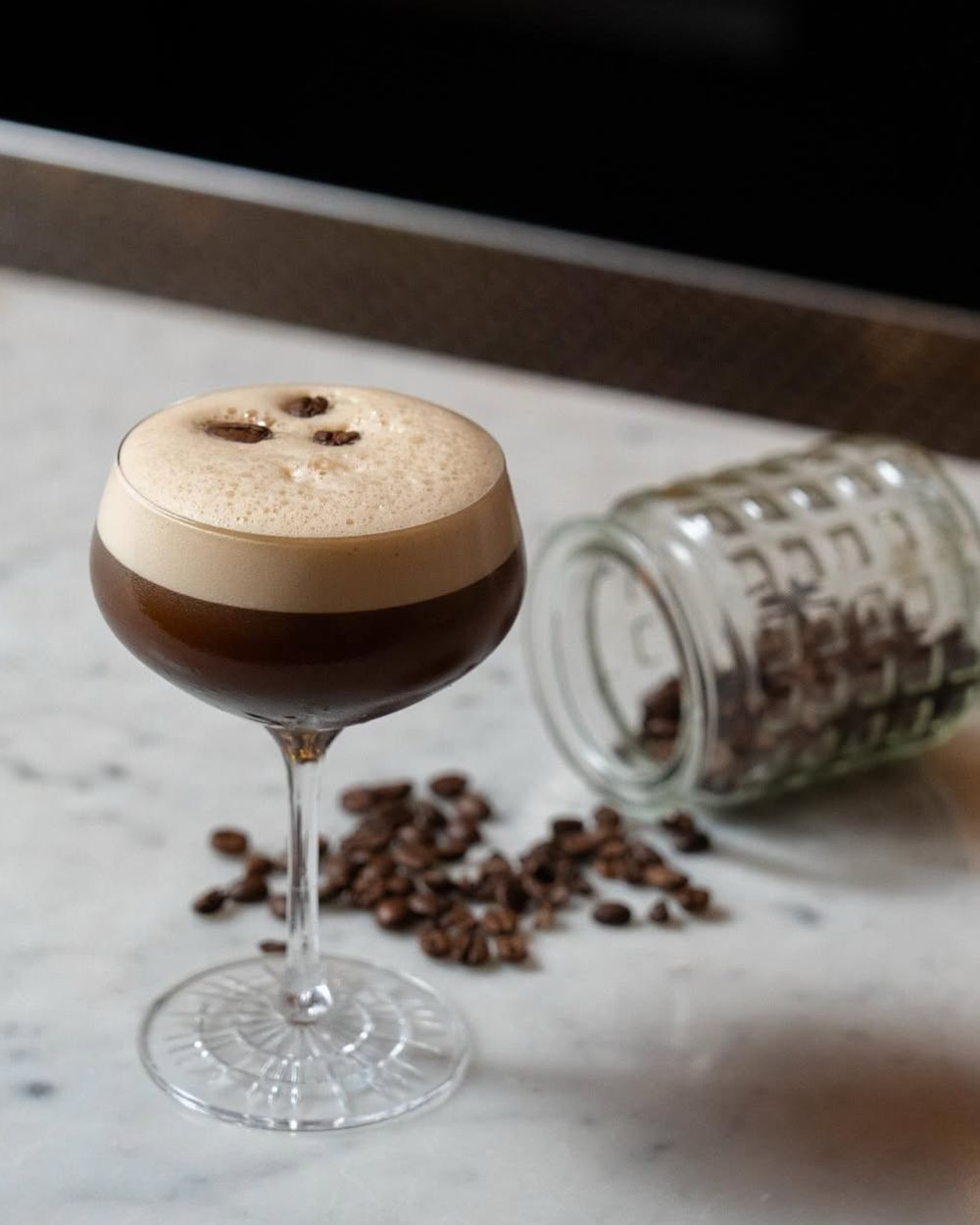 Cocktail glass with frothy dark coffee drink garnished with coffee beans, next to spilled glass jar of coffee beans.